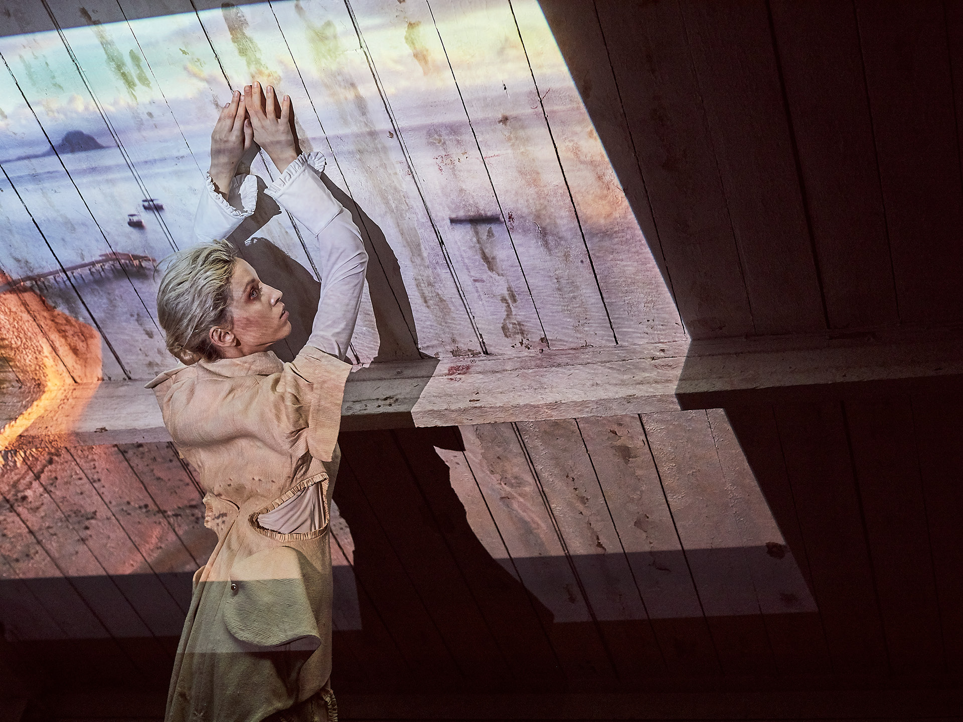 Scandinavian looking white model wearing white and beige clothing holding her hands up against a rough wooden angled ceiling shot by Alex James