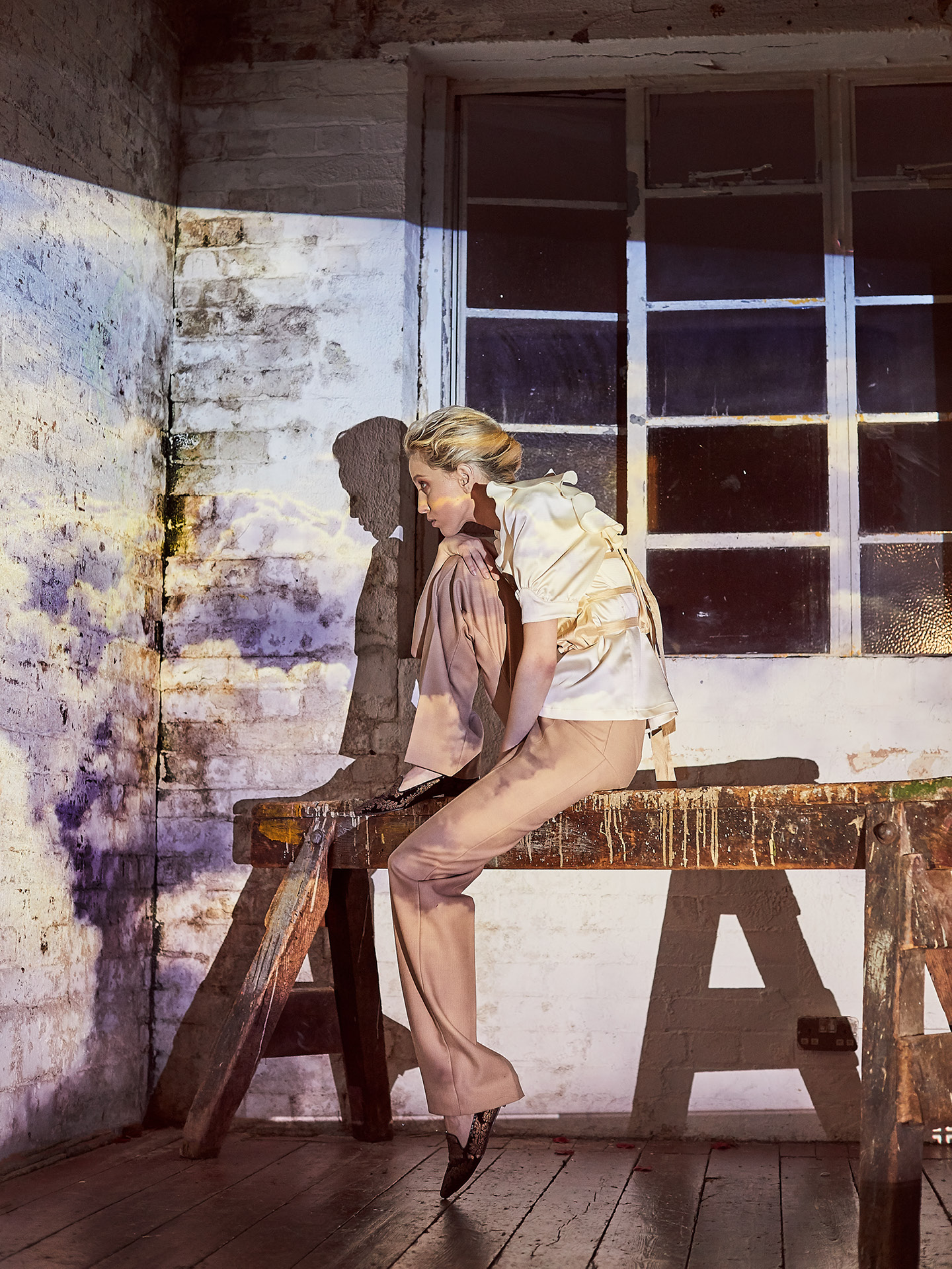 Scandinavian looking white model wearing white and beige clothing sitting on an a-frame near a window and rough white painted brick wall shot by Alex James