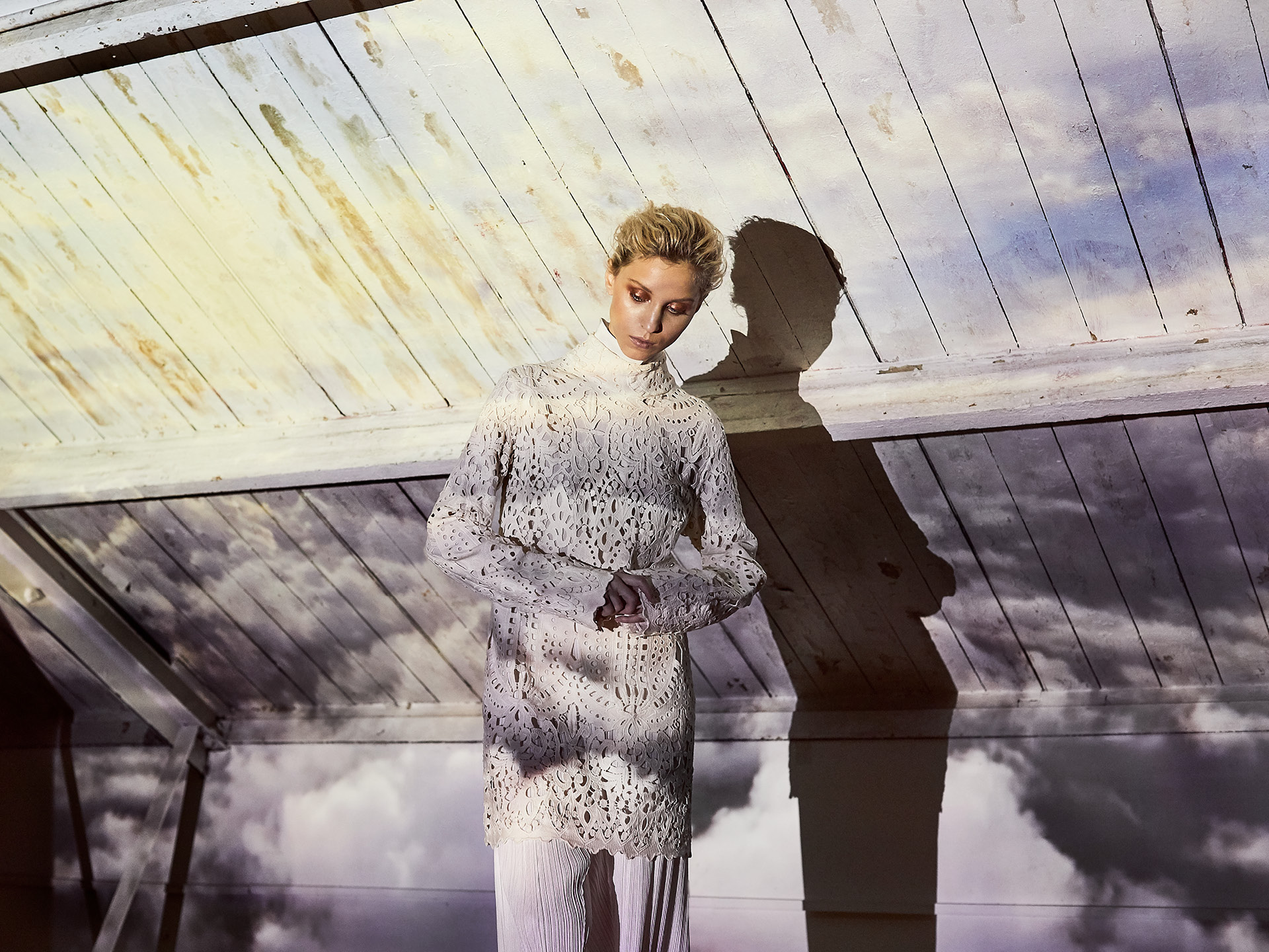 Scandinavian looking white model wearing white and beige clothing standing hunched beneath a slanted wooden ceiling