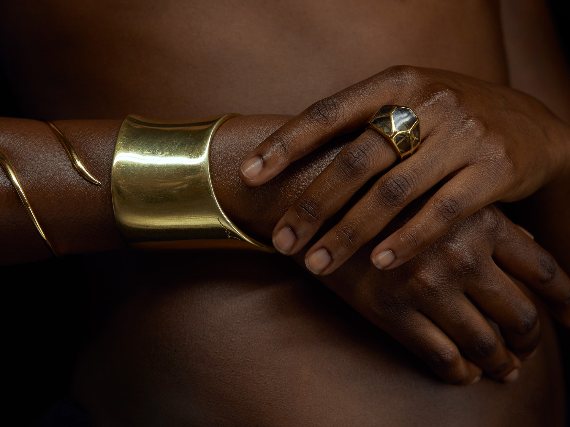 Detailed crop of a black woman’s arms wearing a golden bangle and bracelet with a golden jewelled ring on her hand