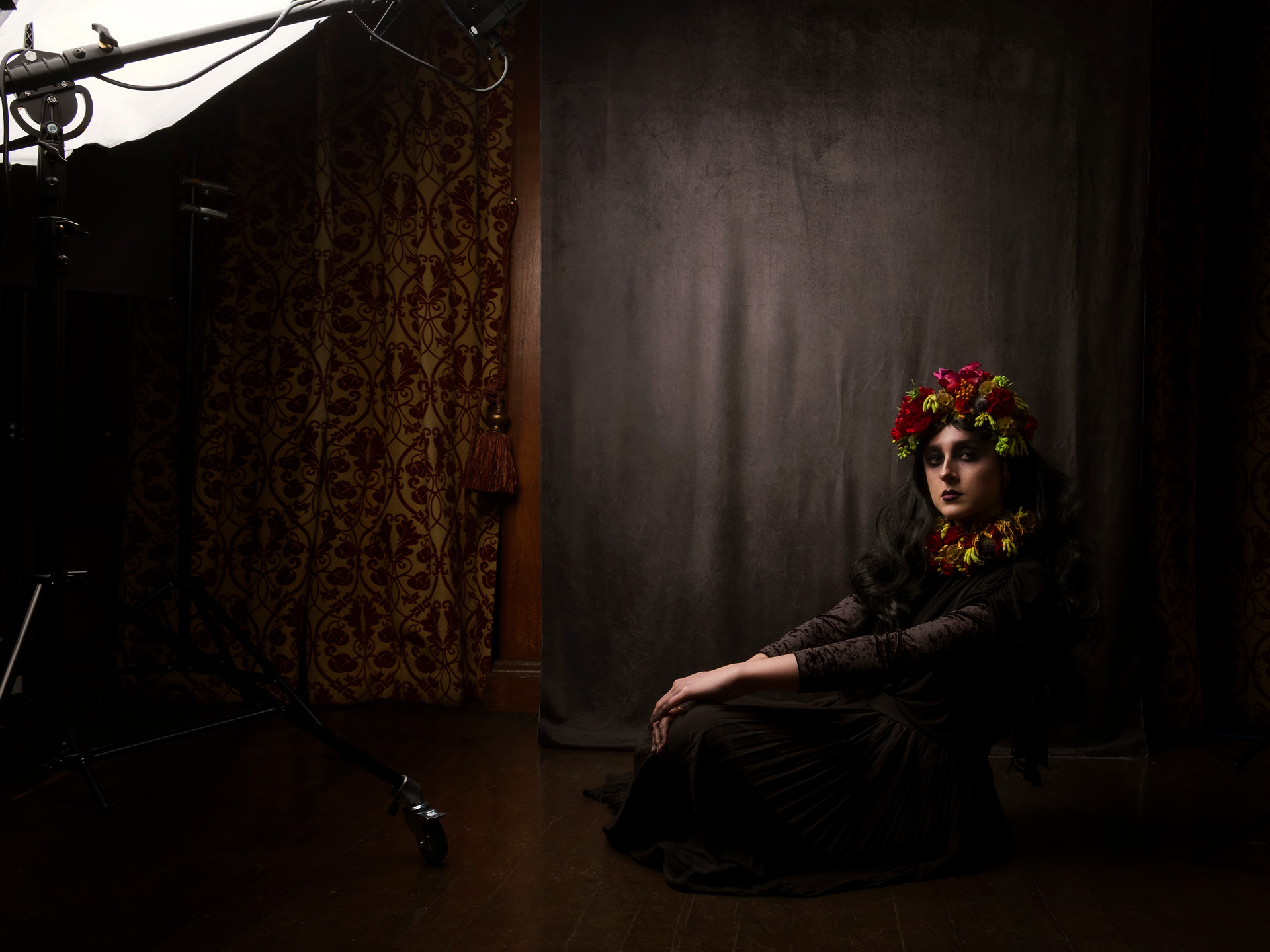 A dark photograph of a model with black hair and deep eyes sitting on the floor with notable background elements of the room still visible like the curtain and lighting gear 