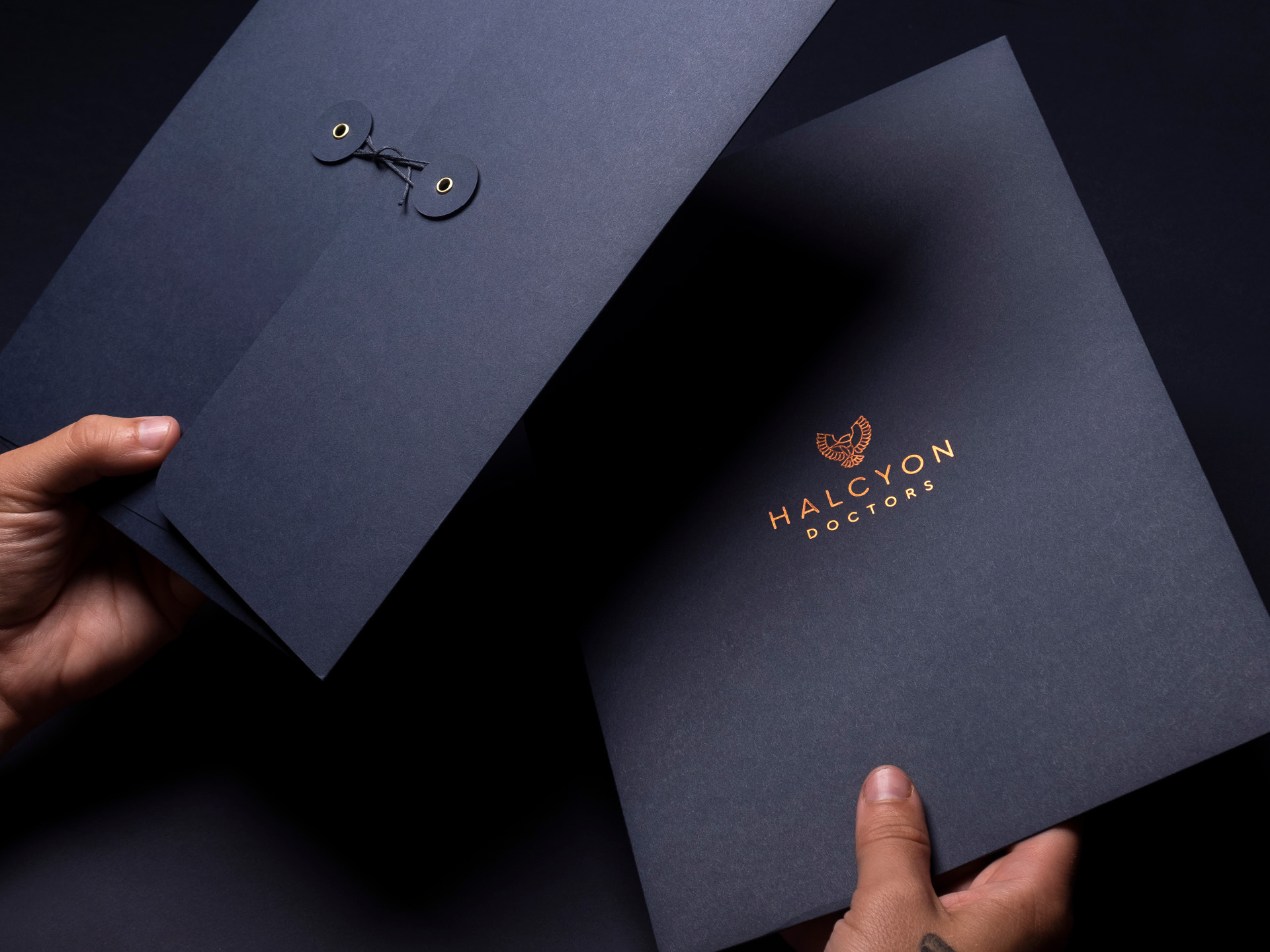 Hands holding two halcyon doctors branded folders with a string and washer binding on one side and the logo copper foiled on the other